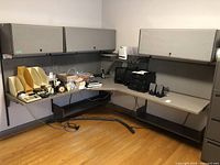 Full view corner office desk with various office items on surface, two wall-mounted cabinets above desk, desk is in L shape with metal legs at ends.