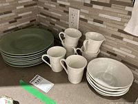 Photo showing stacked green dinner plates, white mugs, and white cereal bowls on a kitchen counter.