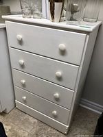 Full view of the white painted wood dresser showing four drawers and round knobs, with contents on top, visible wear on finish.