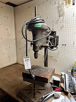 Front angle view of vintage steel drill press showing motor housing, cord wrapped around the top, and rectangular worktable with mounting slots.