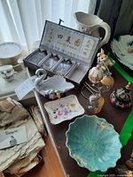 Overview image showing various ceramic and decorative items including hanging egg, bunny figurine, white pitcher, swan ceramic, three perfume jars, floral plates, and boxed gifts.