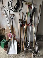 Full view of various garden tools arranged against a wall including shovel, fork, axe, and other hand tools with wooden handles.