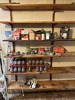 Four basement shelves containing assorted hardware items such as boxes of screws and nails, jars with fasteners, a metal safety check valve, and organizers on wooden shelves.