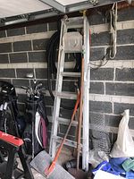 Photo showing full view of the folded 6-foot aluminum ladder, metal snow shovel with orange handle, and wooden handled ice chipper leaning against a wall in a garage.