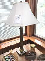 Photo showing the lamp turned off, highlighting the lamp's metal ribbed base, square metallic foot, and white fabric lampshade. Surrounding window frame and some small items on table visible.