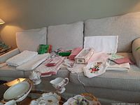 Overall lot showing linens including folded tablecloths, napkins with embroidery, and accessories displayed on a sofa and glass table.