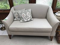 Frontal view of beige upholstered loveseat with curved back, armrests, and two patterned throw pillows on a tiled floor near window.