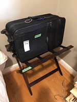 Black soft-sided suitcase placed on wooden luggage stand with straps in corner of room.