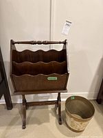 Front and angled view of wooden magazine rack and painted wastebasket placed next to each other on carpeted floor against white wall.