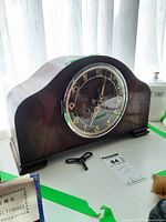 Front view of wooden mantel clock with curved top, displaying glass-covered circular face with gold Arabic numerals and black hands with a visible winding key below.