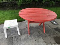 Two tables outdoors on paving stones near grass: round red painted wood table with surface wear and square white plastic side table with some dirt.