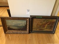 Two vintage framed paintings leaning against a wall on hardwood floor, showing ornate carved frames and landscape scenes.