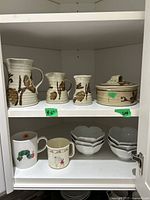 Cabinet shelf with three pottery pitchers and covered bowl with brown leaf design, all ceramic. Lower shelf contains two character mugs and six lotus-shaped ceramic bowls in two stacks.