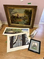 Framed vintage painting with ornate gold frame leaning against a wall, surrounded by several unframed prints laid on the floor including a winter birds print, a water and greenery print, a small narrow Eiffel Tower print, and a small colorful framed illustration.