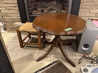 Wooden side table with scalloped edge top and wooden footstool with woven top positioned near a fireplace.