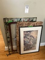 Three framed art pieces stacked leaning against a wall: a stained glass panel, Mexican art, and Asian print.