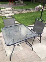 Outdoor square table with metal base and textured glass top next to two grey mesh metal frame folding chairs on patio.