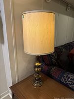 Full view of the lamp showing the fabric shade and the brass base placed near a wall and sofa.