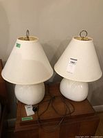 Pair of white ceramic table lamps on a wooden surface showing the bases and shades with electrical cords visible.