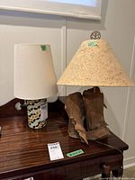 Two table lamps side by side on a wooden table. The lamp on the right has a driftwood base and paper shade, the lamp on the left has a ceramic base with leaf pattern and a fabric shade.