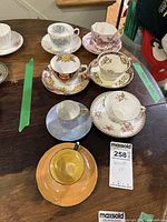 Overview of seven teacups and saucers in various floral and decorative patterns, arranged on wooden surface.