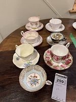 Photo showing six assorted vintage teacups with matching saucers, various floral and gold rim designs on a wooden table.