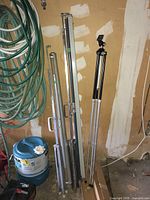 Three metal projector screen stands and a tripod stand with telescoping legs positioned against a wall seen with garden hoses and paint container nearby.