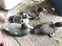 Photo showing all four vintage wooden duck decoys positioned on a rough surface, each with ropes tied around their necks.