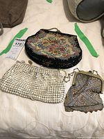 Three vintage evening bags displayed on a white surface: floral tapestry bag, white metal mesh bag, multicolored rhinestone mesh bag.