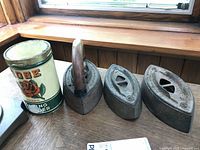 Three vintage cast iron clothes irons on a wooden surface next to a can of washing powder, showing shape, handle, and size.