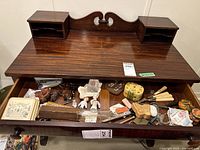 Contents of an open wooden desk drawer showing various vintage small items including trinket boxes, pincushion dolls, tools, and accessories.