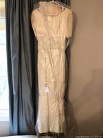 Full view of the vintage wedding dress hung on a hanger beside a window with grey curtains, showing the full length and fitted silhouette under a clear plastic cover.