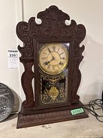 Full view of the wooden mantel clock with ornate carved wood frame and base, golden decorative trim on clock face, and pendulum visible through glass panel.