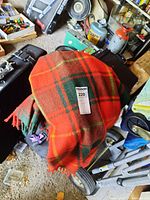 Red and green plaid wool throw blanket spread partially on a black soft seat with clutter in the background.