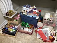 Wide view of multiple boxes, bins, and containers filled with assorted gift wrapping materials including bows, ribbons, wrapping paper, and gift bags on a floor.