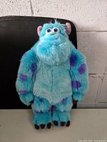 Front view of the blue and purple spotted Sully plush from Monsters Inc. standing on a black surface against a concrete wall.