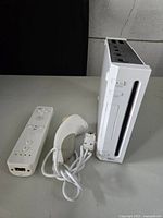 Front view of Nintendo Wii console standing upright with third-party white Wii remote and nunchuck controller laid flat on table.