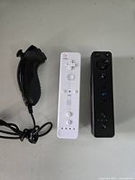 Photo showing a white Wii Remote controller, a black Wii Remote controller, and a black Nunchuck controller, all compatible with Nintendo Wii system.