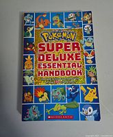 Front cover of the Pokemon Super Deluxe Essential Handbook showing multiple Pokemon characters around a blue border with bold title text in the center.