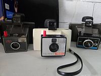 Three vintage Polaroid Land Cameras displayed side by side, showing front view including lenses, flash units, and viewfinders.