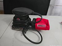 Photo of a vintage Jobmate branded black palm sander with an attached power cord, sitting on a table. The sander has a red dust collection bag also branded Jobmate attached at the rear.