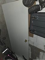 White wooden interior door leaning against a wall with brass-colored doorknob featuring a lock. The door is flat and plain with no panels or windows.