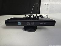 Front view of the Xbox 360 Kinect Sensor showing the three sensor lenses, Xbox 360 branding, cable, and stand base.