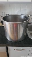 Front angled view of the large stainless steel stock pot placed on a black counter near a kitchen sink, showing the pot's exterior surface, double riveted handles, and open top.