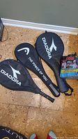 Three Diadora-branded tennis racket protective cases on floor with a pack of Penn tennis balls nearby.