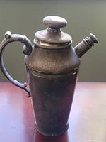 Side view of the antique silver-plated cocktail shaker showing the elegantly curved handle, tapered body, and spout with cap.