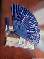 Photo of three handheld fans, one opened blue fan with a floral design and tassel, two others still wrapped in plastic in cream and pink colors.