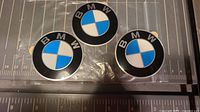 Three BMW badges with black and blue and white colors, each 57 mm diameter, placed on a clear plastic surface with measuring scale underneath.