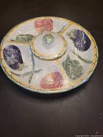 Top view of a round ceramic compartmented serving platter with four sections and a removable lid with a knob, decorated with colorful vegetable motifs.