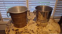Two metal champagne buckets side by side on a granite counter under window blinds. Left bucket has a brushed matte finish and ring handles, right bucket has a polished reflective finish and missing one handle.